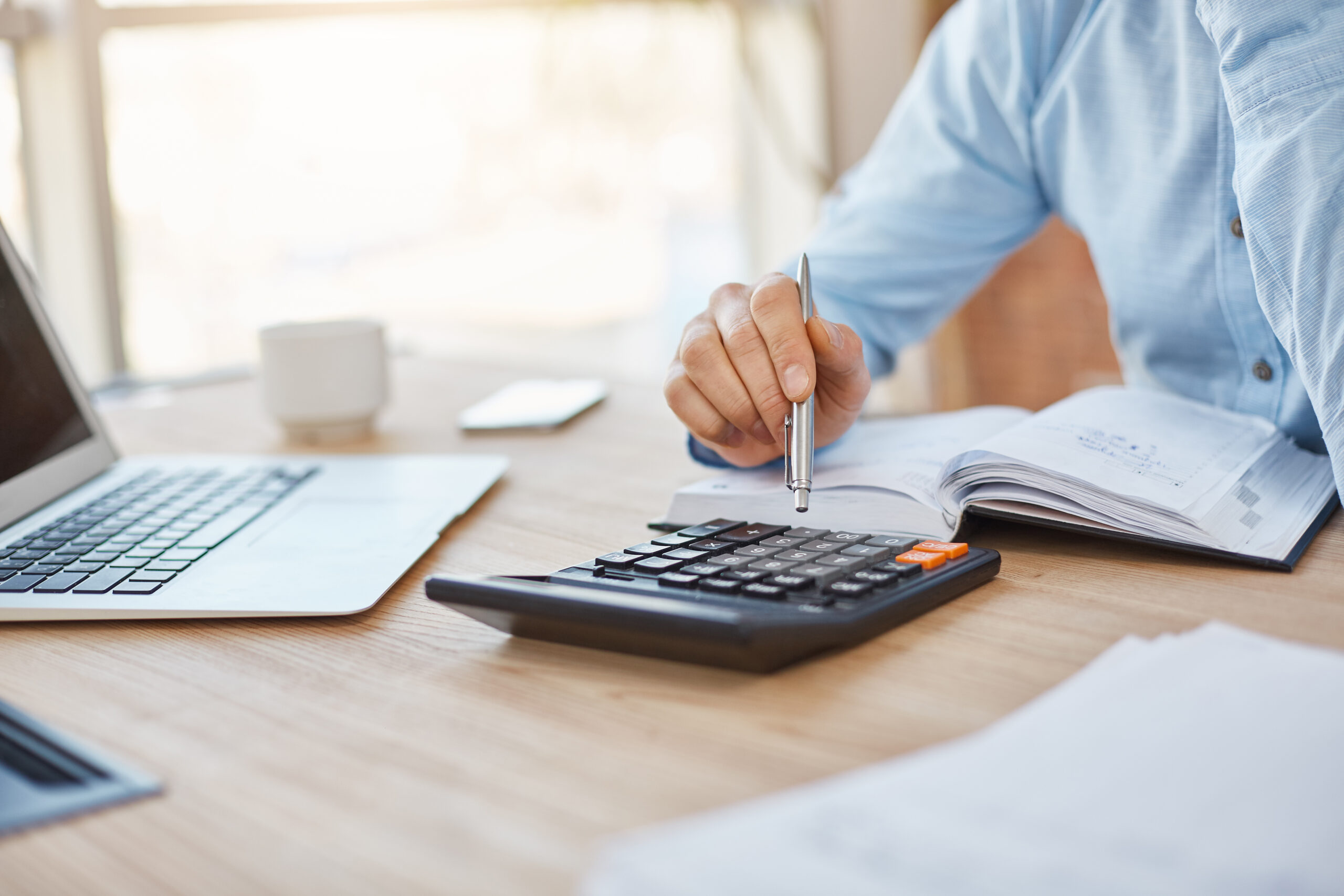 Close up detail of professional serious accountant sitting in light office, checking company finance profits on calculator, writing down results in notebook