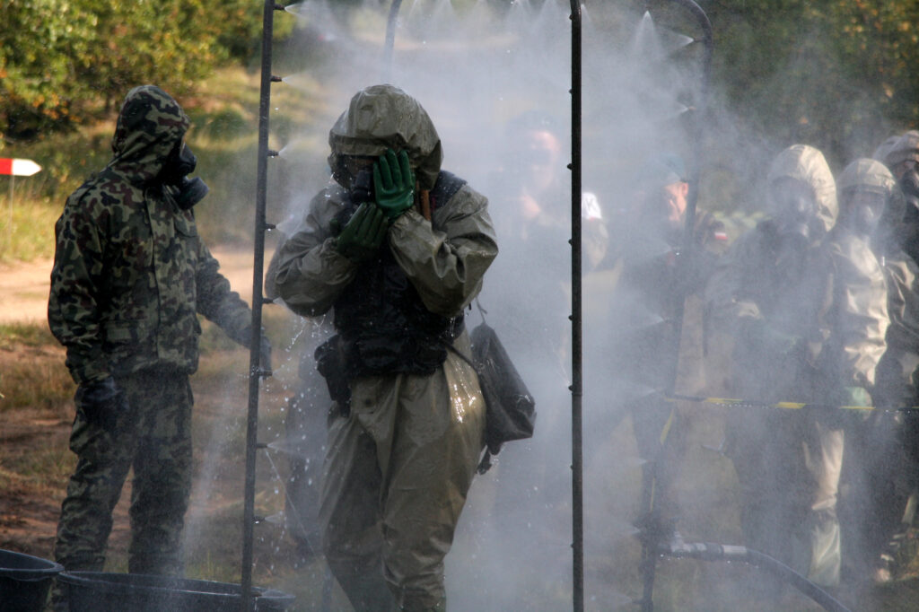 Military personnel wearing hazmat suits and gas masks move through a decontamination spray tent during a training exercise