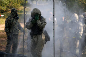Military personnel wearing hazmat suits and gas masks move through a decontamination spray tent during a training exercise
