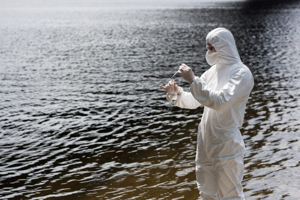 Water inspector in protective costume, latex gloves and respirator taking water sample at river